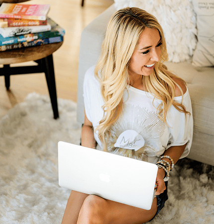 Danette sitting on a rug with an open laptop, smiling as she reads.
